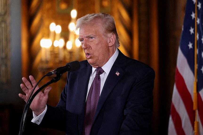 Trump speaking at a microphone in an opulent gold room with a US flag behind him