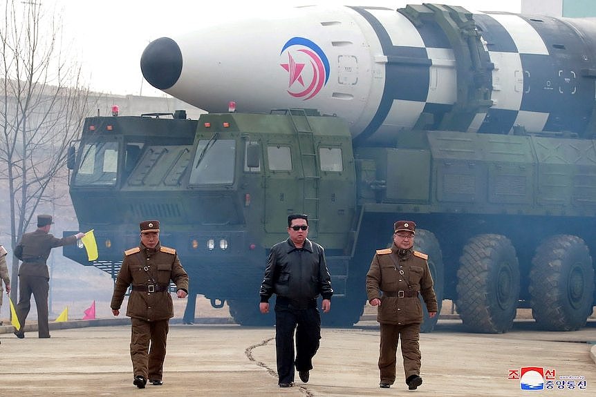 Kim Jong Un and two uniformed military sidekicks walk away from a large missile lying on a vehcile. 