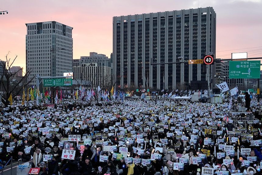 A huge crowd of protesters, many holding signs, rally in an open space to demand the arrest of impeached South Korean president.