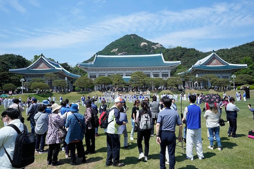 A large crowd gathers on the large front lawn of an Oriental-looking building with a blue roof