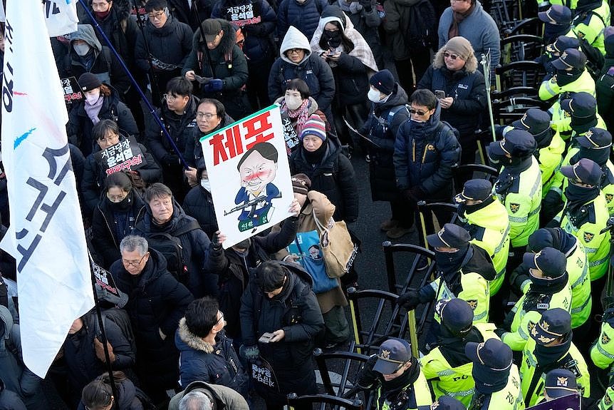 Protesters hold signs satirising impeached South Korean President Yoon Suk Yeol near a barricade behind which police wait.