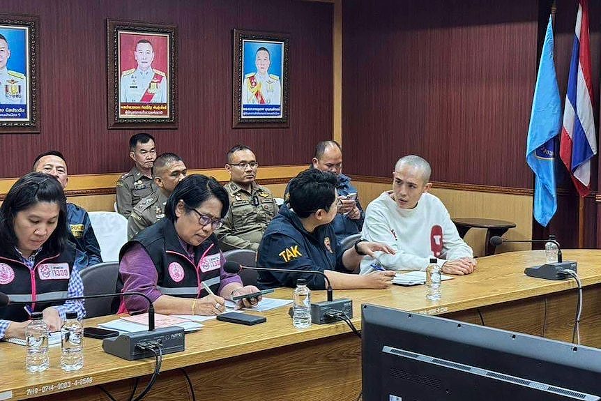 Wang Xing wearing a white jumper sitting alongside Thai police officers in dark and green jumpsuits 