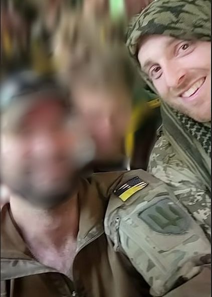 Two men in khaki army gear smile in a selfie, with one's face blurred.