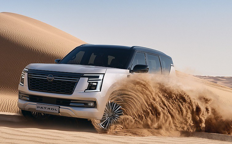 The all-new Nissan Patrol with white body is cruising through the desert, kicking up sand.