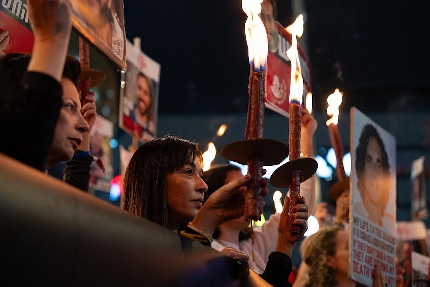 a woman in a crowd holds a torch amid signs in Hebrew, looking solemn