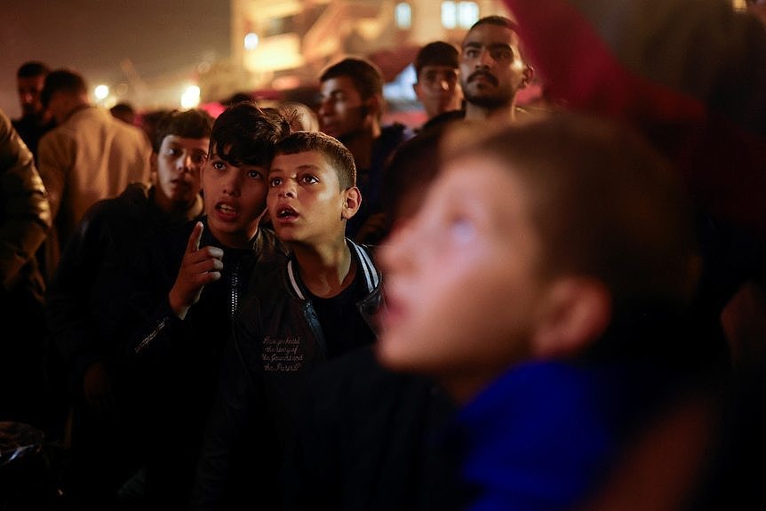 Two Palestinian children in a public crowd looking up at a large screen showing the announcement of a ceasefire.