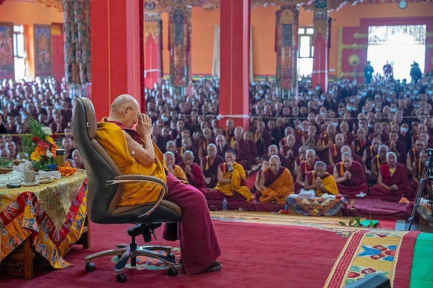 The Dalai Lama is seated before a large crowd as he leads prayers at a lavish golden monastery.