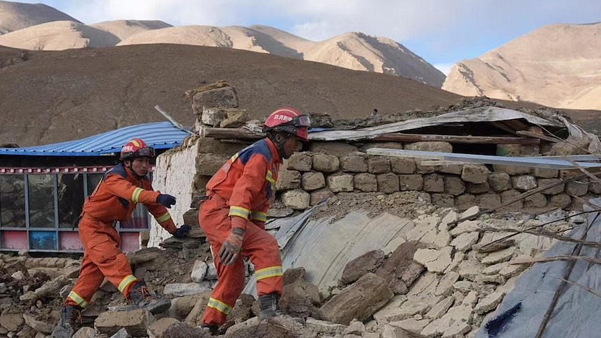 Two rescue workers in orange suits and helmets search rubble for survivors after an earthquake in a mountainous region.