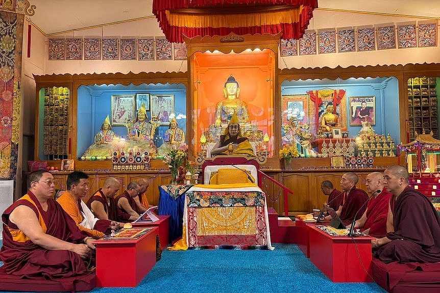 A row of five robed monks sit opposite a row of four robed monks in a small and bright buddhist temple.