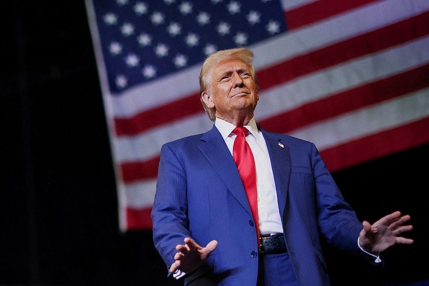 Trump is viewed from below at a campaign rally, in front of an American flag.