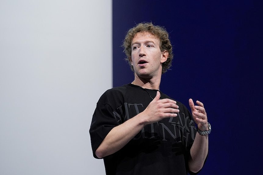 Mark Zuckerberg, standing on a stage, wearing a black t-shirt, talking at a company conference.