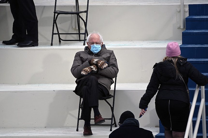  Senator Bernie Sanders sits on a fold=up chair wearing a blue face mask, a grey jacket and brown, white and black mittens