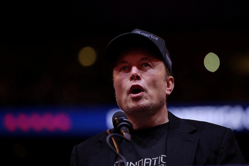 Elon Musk wears a black cap as he speaks on stage 