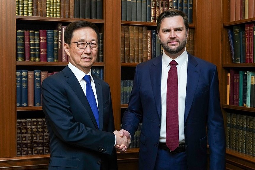 Han Zheng and JD Vance shake hands while standing in front of a wooden bookcase of leather-bound books.