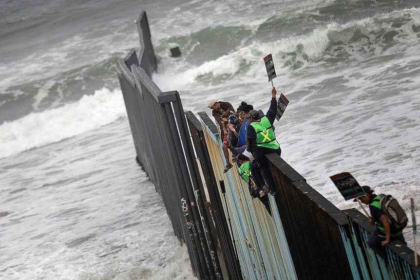 Migrants sit on top of the border wall on the beach, with waves crashing underneath them.