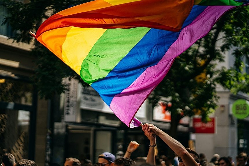 Someone waves a rainbow coloured flag in the sunlight above a crowd of people