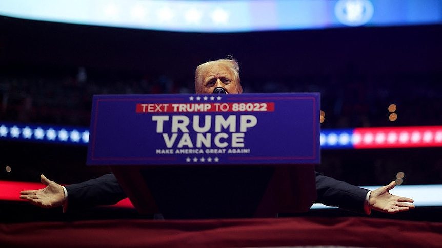 Donald Trump stands behind a podium with his arms open. A Trump-Vance campaign placard is attached to the podium.