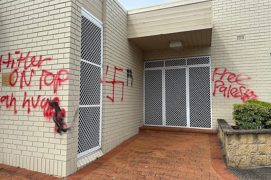 Anti-Semitic graffiti mentioning Hitler and showing a swastika along a brick wall.