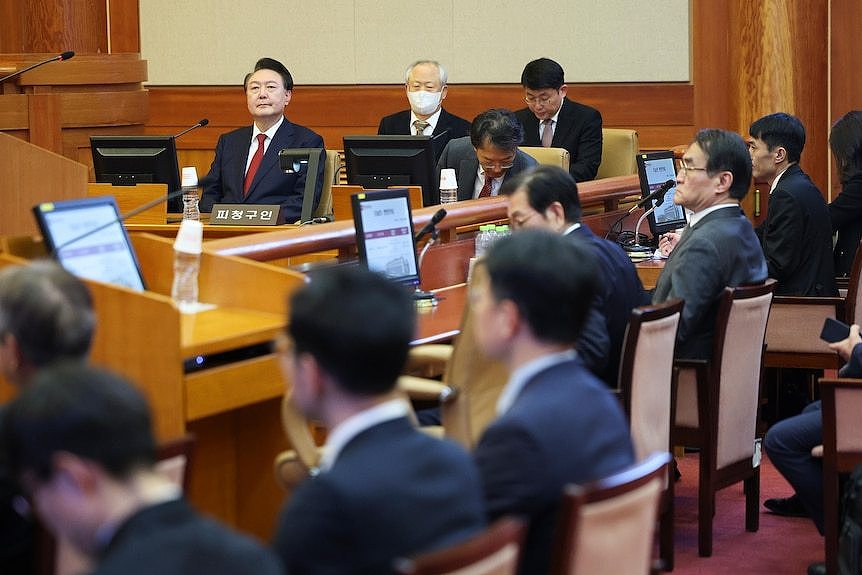 Men in a courtroom sit in suits with some looking at computer screens