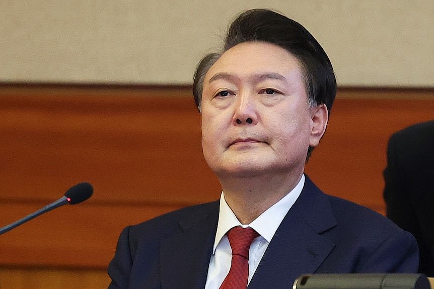 A man with black hair combed back sits in a suit with a red tie behind a microphone looking past the camera