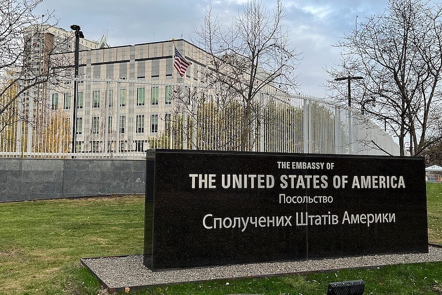 A black stone sign reading 'The United States of America' in English and Ukrainian in front of a silver fence and pale building