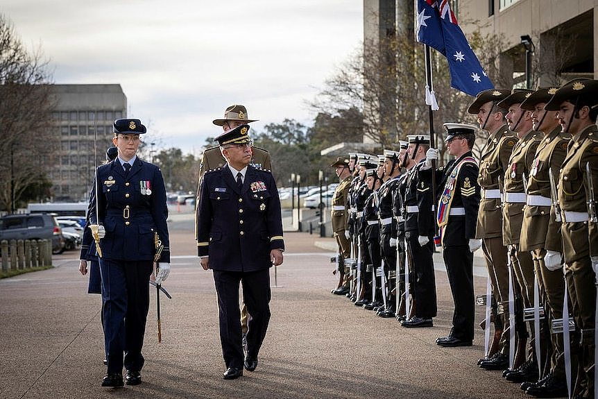 Two people in military uniforms walk past an armed guard. 