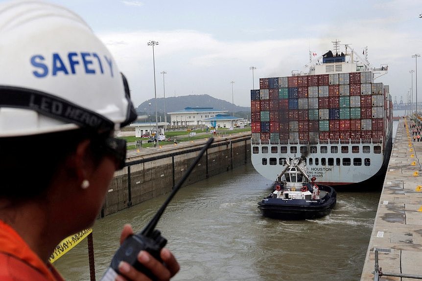 An image of a worker on a walkie-talkie in foreground blurry with large ship in focus in background.