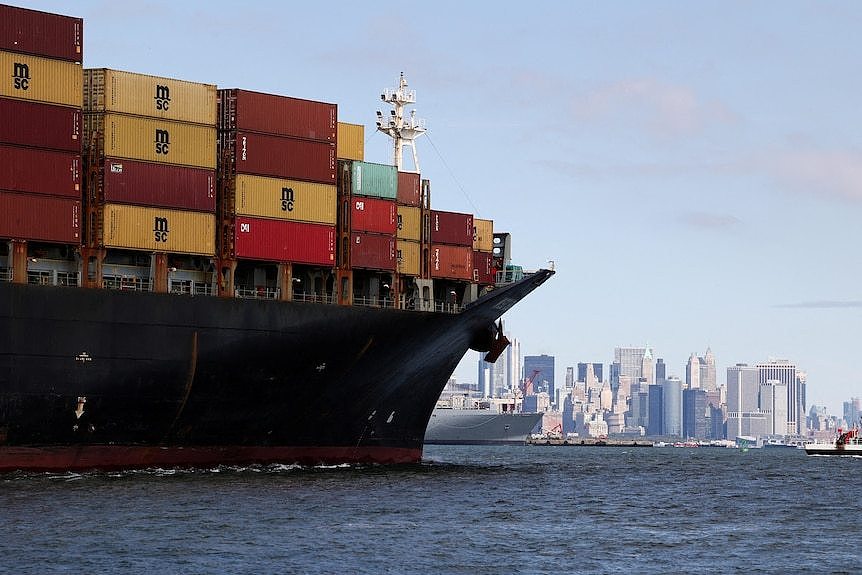 Large colourful shipping containers on board a large ship sailing on a bay. The New York City skyline is in the distance.