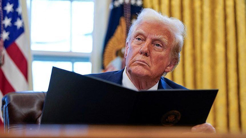 Donald Trump sits at a desk holding a black folder open with yellow-gold curtains and two flags behind him.