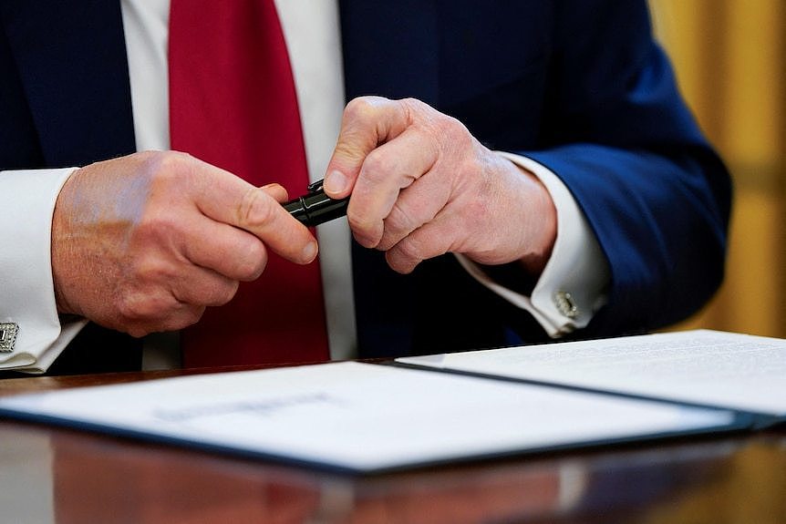 A man wearing a suit and red tie caps a black pen lid onto a marker after signing a document.