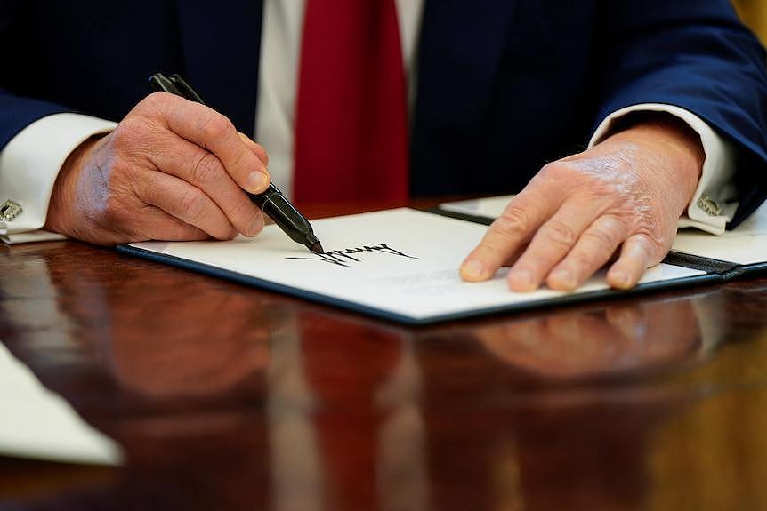 A man uses a black marker to write his signature on a piece of paper inside a black folder on a desk.