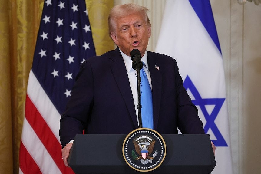 Donald Trump stands at a lectern in front of the US and Israeli flags.