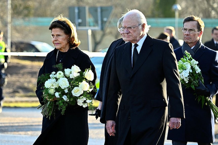 The king and queen, wearing black, laying flowers on the ground outside a school.