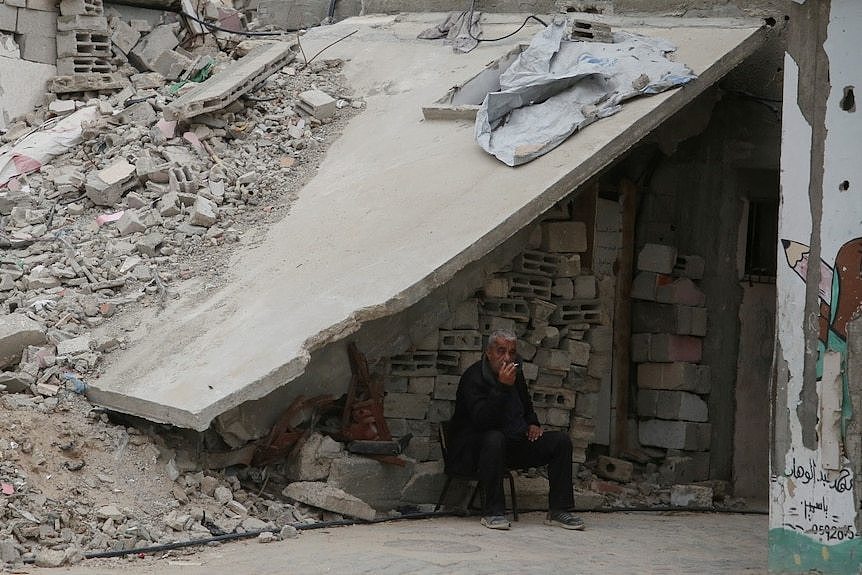 A man sits near the rubble of a building, smoking a cigarette.