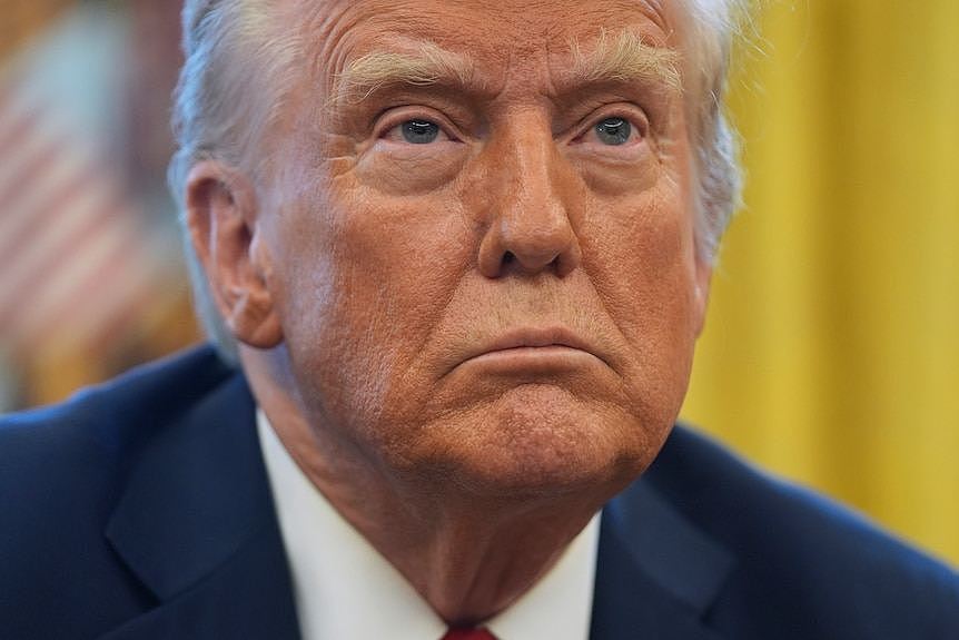 A closeup of Trump looking orange as he listens to someone else speaking