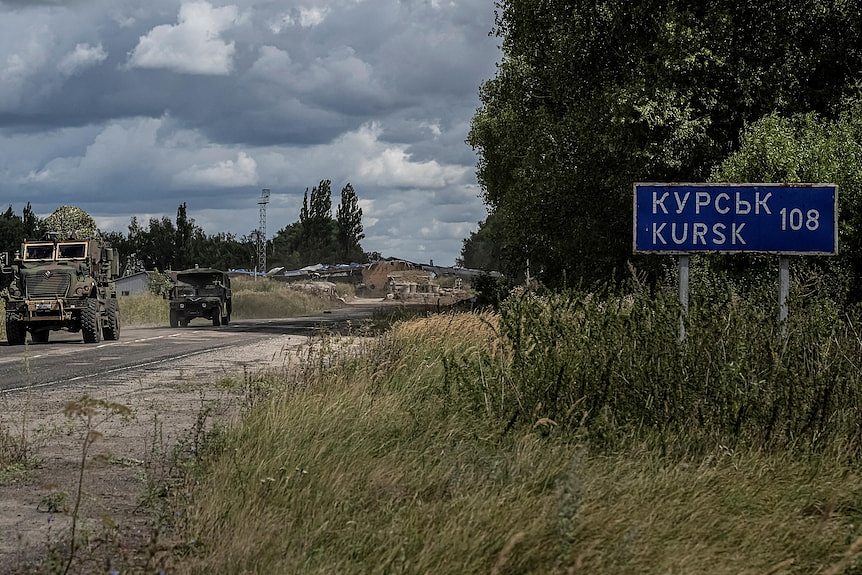 A column of military vehicles driving past a sign saying there's 108 kilometres to Kursk.