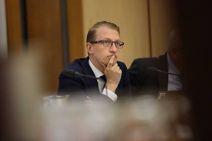 A man in a suit and glasses sits with his hand on his chin at a Senate Committee.