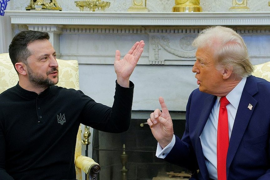 A man in a black jumper and another man in a blue suit and red tie speak and gesture to each other