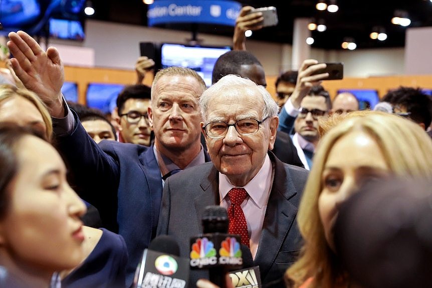 An older man wearing a suit and glasses walks through a crowd of people holding microphones towards him.