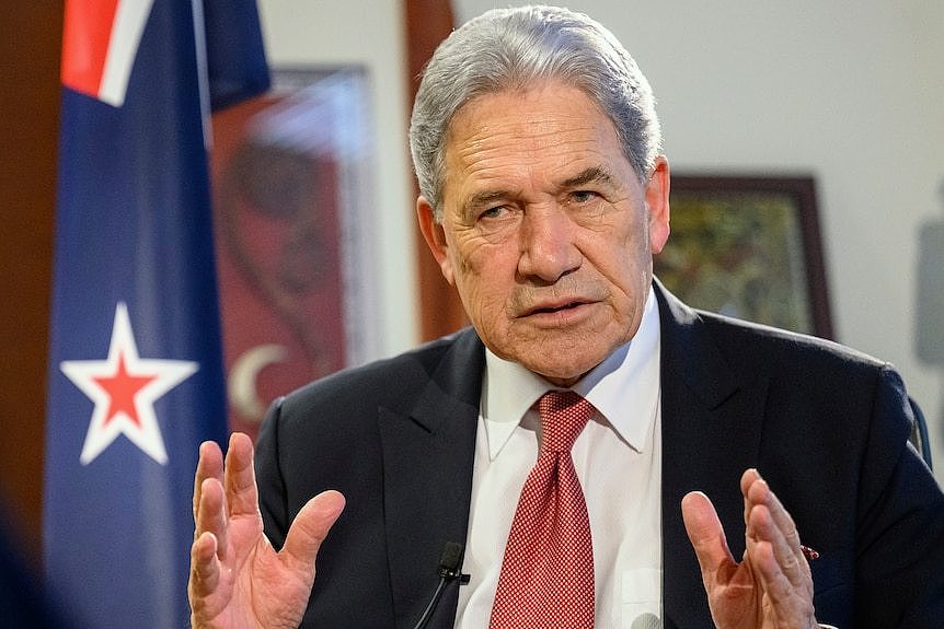 A man with grey hair and in a black jacket and wearing a white shirt and red tie, sitting in front of a New Zealand flag.