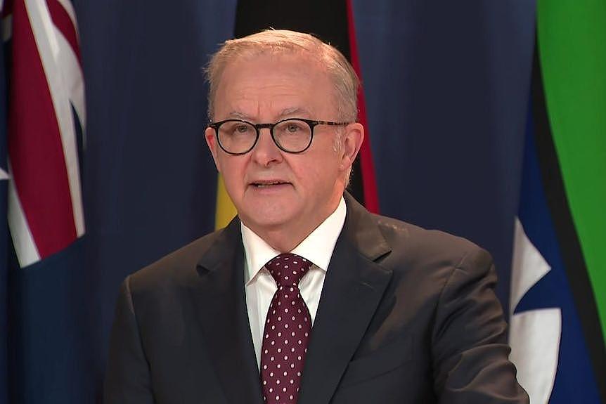 Anthony Albanese wearing glasses speaking at a press conference in front of flags.