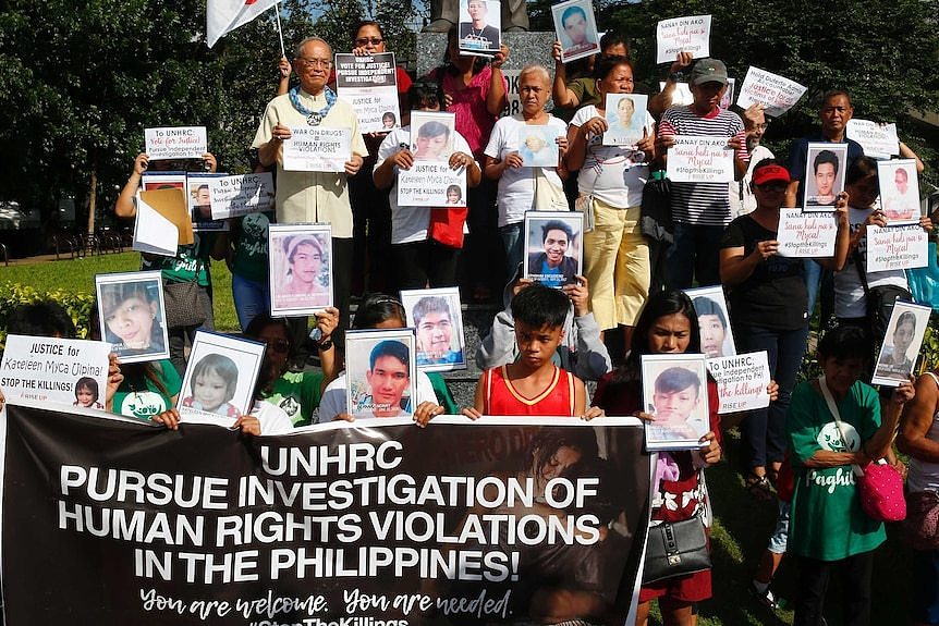 A crowd of people hold placards and portraits of relatives and friends who stand behind a large black banner calling on UN help.