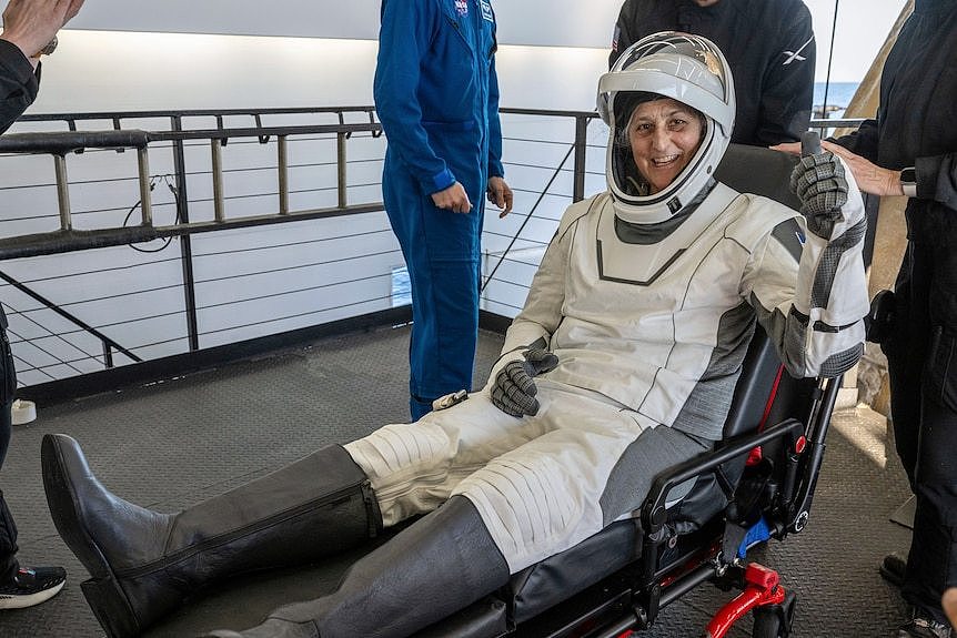 Suni Williams smiles and gives a thumbs up while lying in a stretcher wearing a white padded suit and helmet