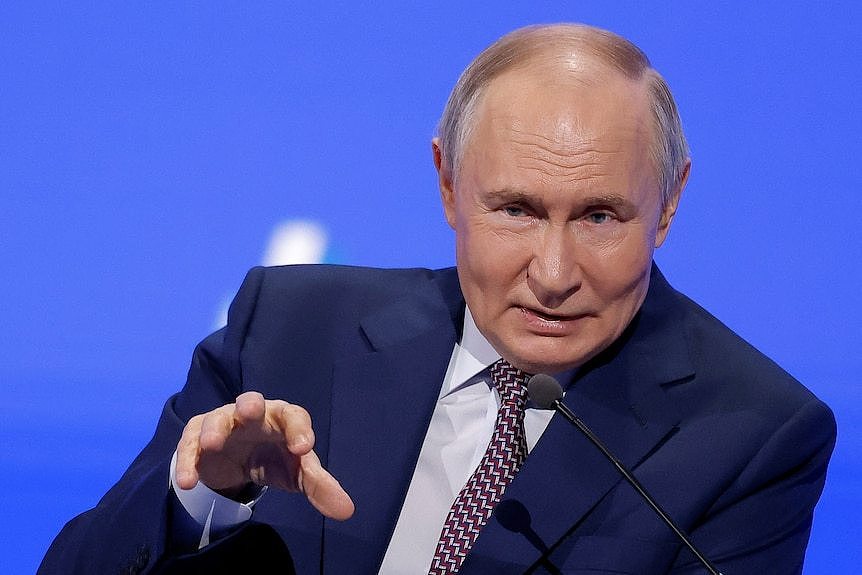 Vladimir Putin, waring a suit and tie and making a hand gesture, pictured in front of a blue background