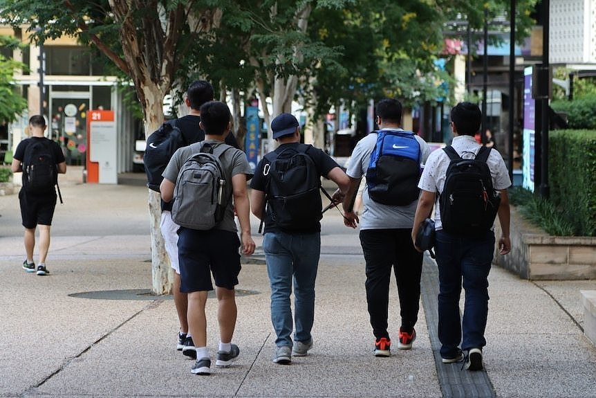 a group of male students wearing backpacks walk through a uni campus away from the camera. their faces can't be seen