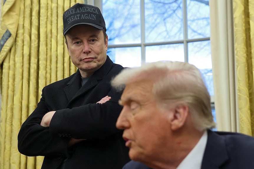 A man wearing all black with a cap stands with his arms crossed looking down at Donald Trump speaking.